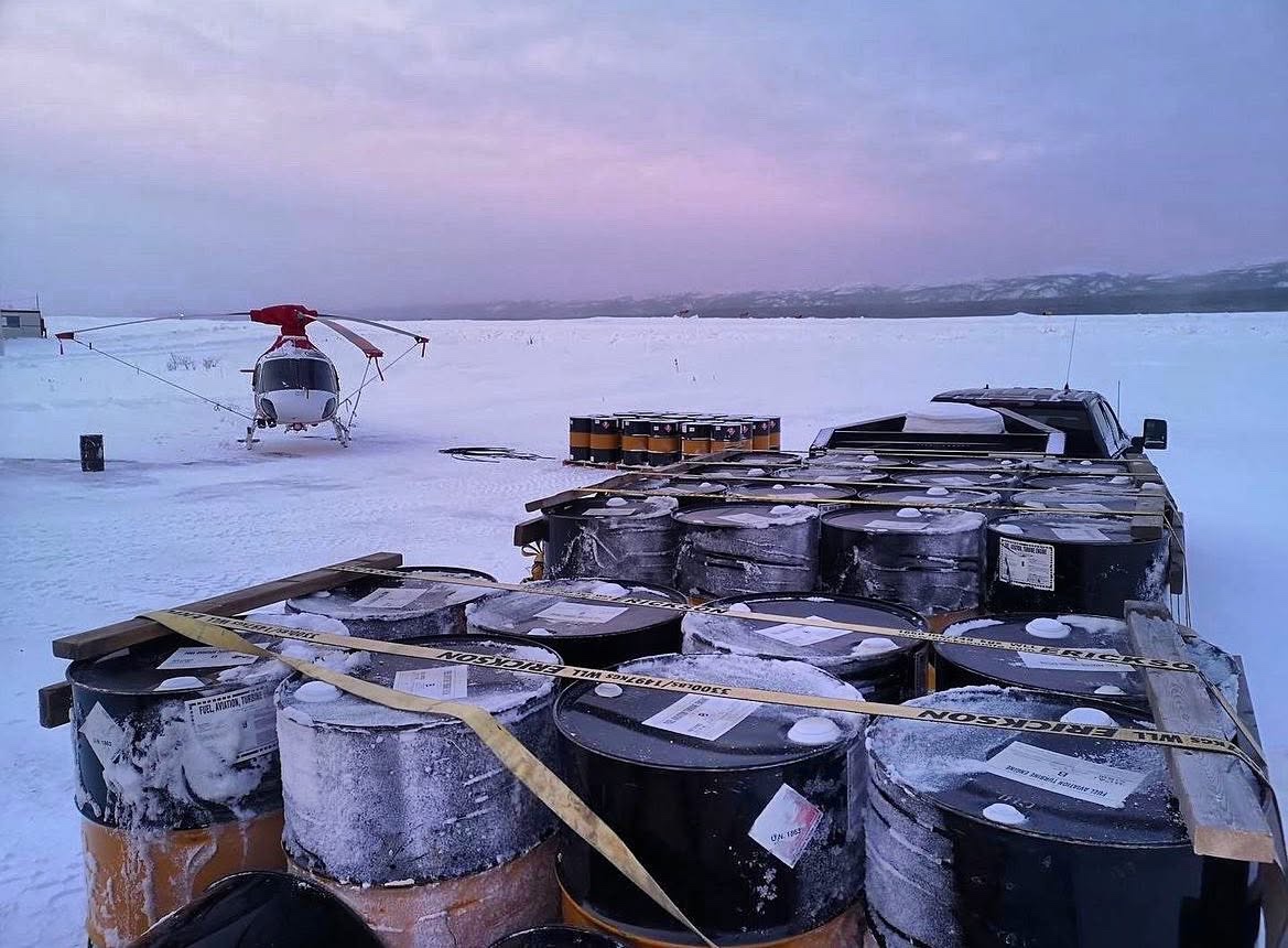 Snow-covered fuel drums secured on a trailer with a helicopter at a remote winter site