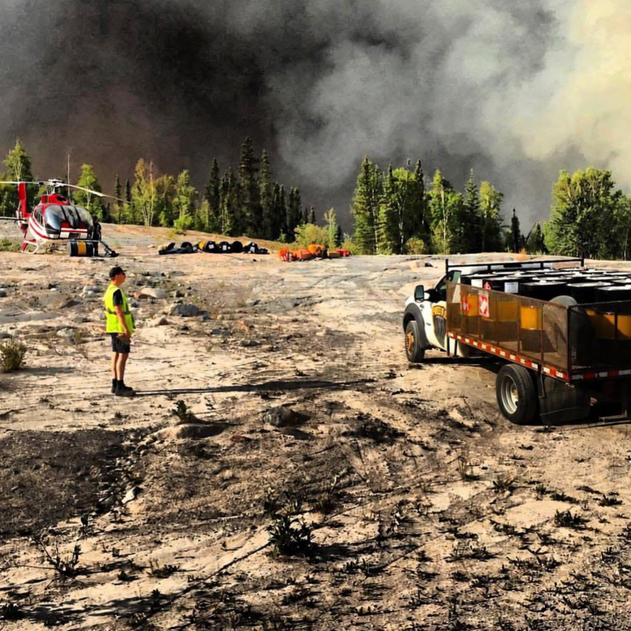 Fuel Flo truck carrying fuel drums beside a helicopter with wildfire smoke behind the trees